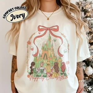 May include: Ivory Comfort Colors t-shirt featuring a castle design with a red bow and the words "Happily Ever After." The design includes illustrations of princesses and fireworks. The shirt is a cream color.
