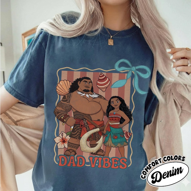 May include: A navy blue t-shirt featuring a graphic of two cartoon characters with the text "DAD VIBES". The design includes a seashell, a hook, and a flower. The shirt has a comfort colors denim label.