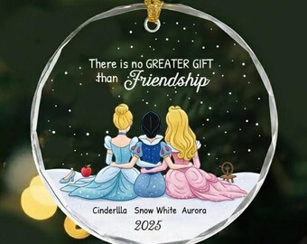Puede incluir: Un adorno de cristal redondo y transparente con tres figuras ilustradas sentadas juntas. El texto dice: There is no GREATER GIFT than Friendship. Debajo de las figuras aparecen los nombres Cinderella, Snow White, Aurora y el año 2025.