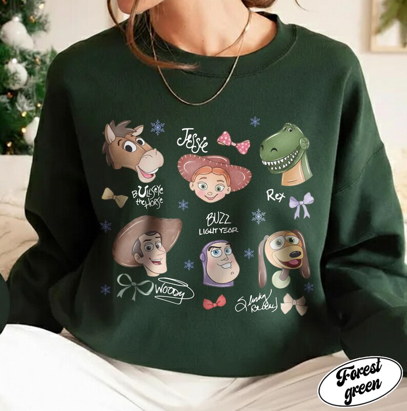 May include: Forest green sweatshirt featuring cartoon character faces from a popular animated movie. The design includes characters like Woody, Buzz Lightyear, and Jessie, with their names and decorative elements. The sweatshirt is a crew neck style.