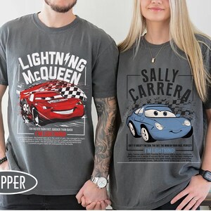 May include: Two gray t-shirts featuring characters from the movie Cars. One shirt displays Lightning McQueen with text, and the other features Sally Carrera with text. Both shirts have a vintage, distressed look.