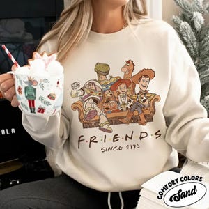 May include: Sand-colored sweatshirt featuring a graphic of characters from the movie Toy Story, seated on a couch. The word "FRIENDS" is printed below the graphic, with "SINCE 1995" underneath. The sweatshirt has a Comfort Colors label.