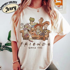 May include: Ivory-colored t-shirt featuring a graphic of Toy Story characters sitting on a couch. The text "FRIENDS SINCE 1995" is printed below the graphic. The shirt has the words "COMFORT COLORS" and "Ivory" printed on the top left.
