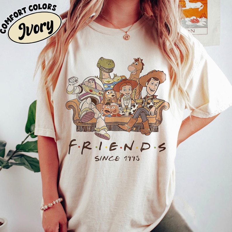 May include: Ivory-colored t-shirt featuring a graphic of Toy Story characters sitting on a couch. The text "FRIENDS SINCE 1995" is printed below the graphic. The shirt has the words "COMFORT COLORS" and "Ivory" printed on the top left.