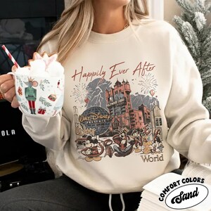 Puede incluir: Sudadera de color arena con un gr&aacute;fico que presenta el texto "Happily Ever After" y personajes de Disney. El dise&ntilde;o incluye el Hollywood Tower Hotel, fuegos artificiales y el logotipo de Walt Disney World. La sudadera es de Comfort Colors.