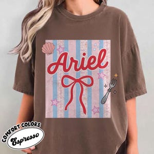 May include: Espresso-colored t-shirt featuring a graphic of Ariel with a bow, stars, and a fork. The design includes a seashell and is set against a pink and blue striped background. The shirt is part of the Comfort Colors collection.