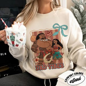 May include: Sand-colored sweatshirt featuring a graphic of two animated characters with the text "DAD VIBES." The design includes a seashell, flower, and a blue bow. The sweatshirt is made of Comfort Colors material.