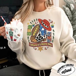 May include: A sand-colored crewneck sweatshirt featuring a graphic print of a cowboy boot, a red hat, alphabet blocks, and small green toy soldiers. The text "WOODY" is on the boot sole, and the text "I Knew It I Knew You." is printed below.