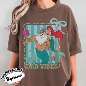 May include: Espresso-colored t-shirt featuring a cartoon graphic of a merman and a mermaid with the text "DAD-VIBES". The graphic is set against a blue and white striped background with a seashell, starfish, and bow.