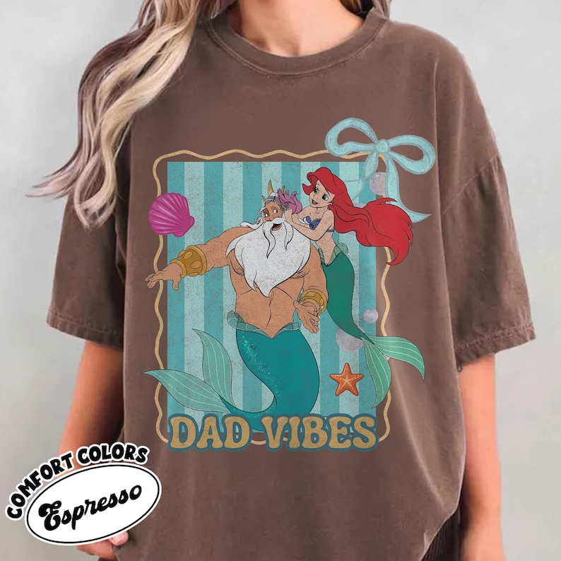 May include: Espresso-colored t-shirt featuring a cartoon graphic of a merman and a mermaid with the text "DAD-VIBES". The graphic is set against a blue and white striped background with a seashell, starfish, and bow.
