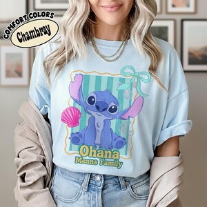 May include: Light blue t-shirt with a cartoon illustration of a blue alien character with large ears and the text "Ohana Means Family". The design includes a pink seashell and a green bow. The shirt is labeled "Comfort Colors Chambray".