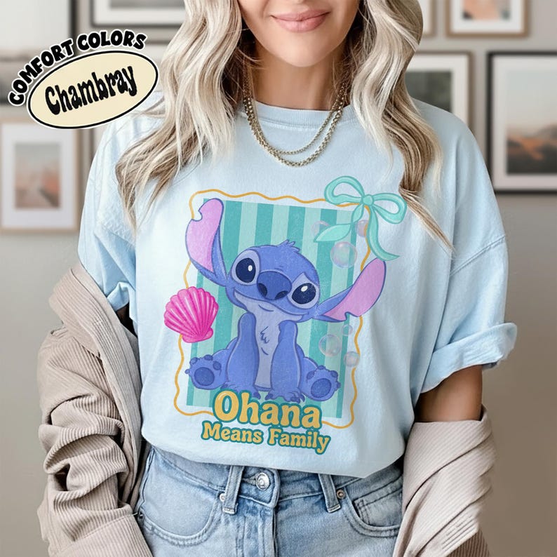 May include: Light blue t-shirt with a cartoon illustration of a blue alien character with large ears and the text "Ohana Means Family". The design includes a pink seashell and a green bow. The shirt is labeled "Comfort Colors Chambray".