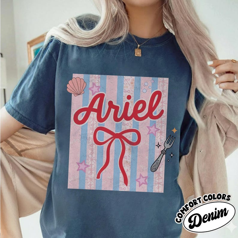 May include: A navy blue t-shirt featuring the name "Ariel" in red script, set against a pink and blue striped background with a bow, seashell, stars, and a fork graphic. The shirt has a "Comfort Colors Denim" logo.