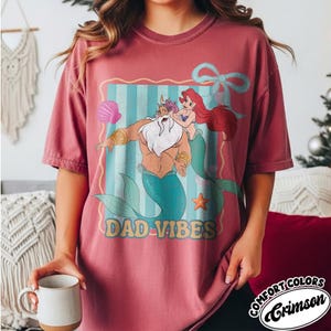 May include: Crimson Comfort Colors t-shirt featuring a cartoon graphic of King Triton and Ariel with the text "DAD VIBES". The graphic is framed with a blue and white striped background and a light blue bow.