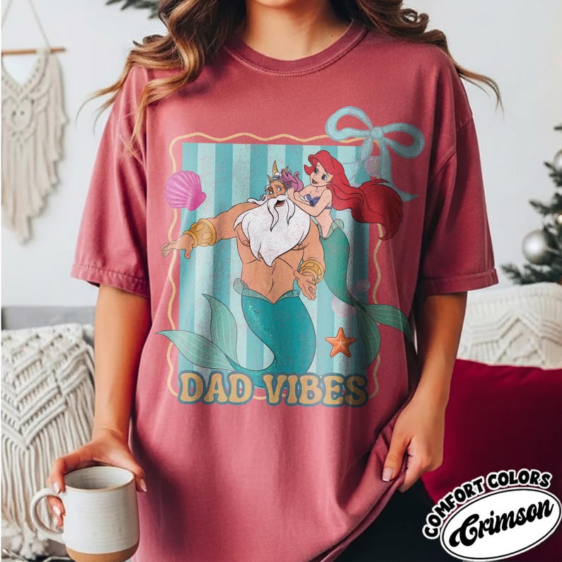 May include: Crimson Comfort Colors t-shirt featuring a cartoon graphic of King Triton and Ariel with the text "DAD VIBES". The graphic is framed with a blue and white striped background and a light blue bow.