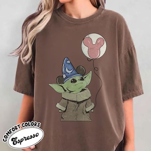 May include: Espresso-colored t-shirt featuring a green alien character wearing a wizard hat with Mickey Mouse ears. The character holds a balloon with a pink Mickey Mouse head design. The shirt has the words "COMFORT COLORS" and "Espresso" in a circular design.
