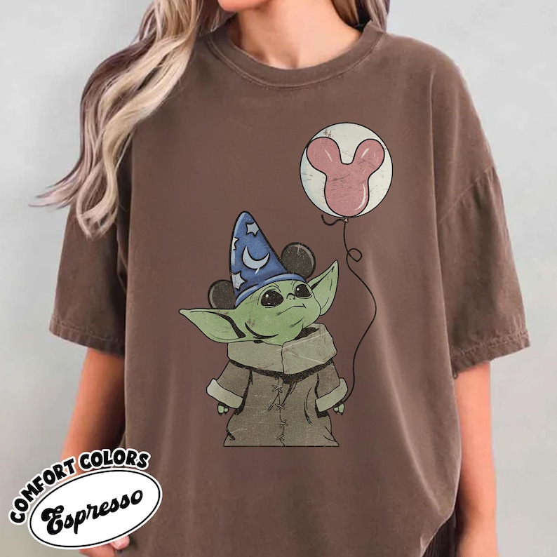 May include: Espresso-colored t-shirt featuring a green alien character wearing a wizard hat with Mickey Mouse ears. The character holds a balloon with a pink Mickey Mouse head design. The shirt has the words "COMFORT COLORS" and "Espresso" in a circular design.