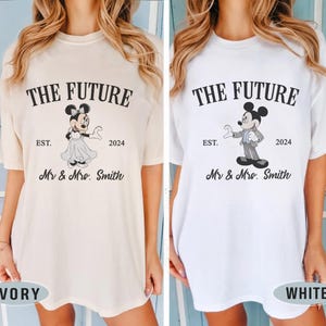 Puede incluir: Dos camisetas blancas con un estampado gr&aacute;fico negro de Mickey y Minnie Mouse. El texto en las camisetas dice "The Future" con el a&ntilde;o "2024" y "Est.". El texto tambi&eacute;n dice "Mr. & Mrs. Smith". Las camisetas se muestran en dos modelos, uno con una camiseta de color marfil y el otro con una camiseta blanca.
