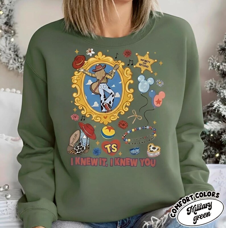 Puede incluir: Una sudadera verde militar con un estampado gr&aacute;fico de una persona con sombrero de vaquero y botas dentro de un marco dorado, rodeada de iconos que incluyen globos, una estrella con el texto INFINITY AND BEYOND y el texto I KNEW IT, I KNEW YOU.