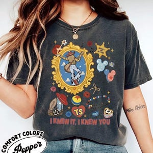 Puede incluir: Una camiseta gris carb&oacute;n con un estampado gr&aacute;fico que presenta un marco dorado ornamentado central con una figura con guitarra, una estrella con el texto INFINITY AND BEYOND, globos, una bota de vaquero, una pelota y el texto I KNEW IT, I KNEW YOU.