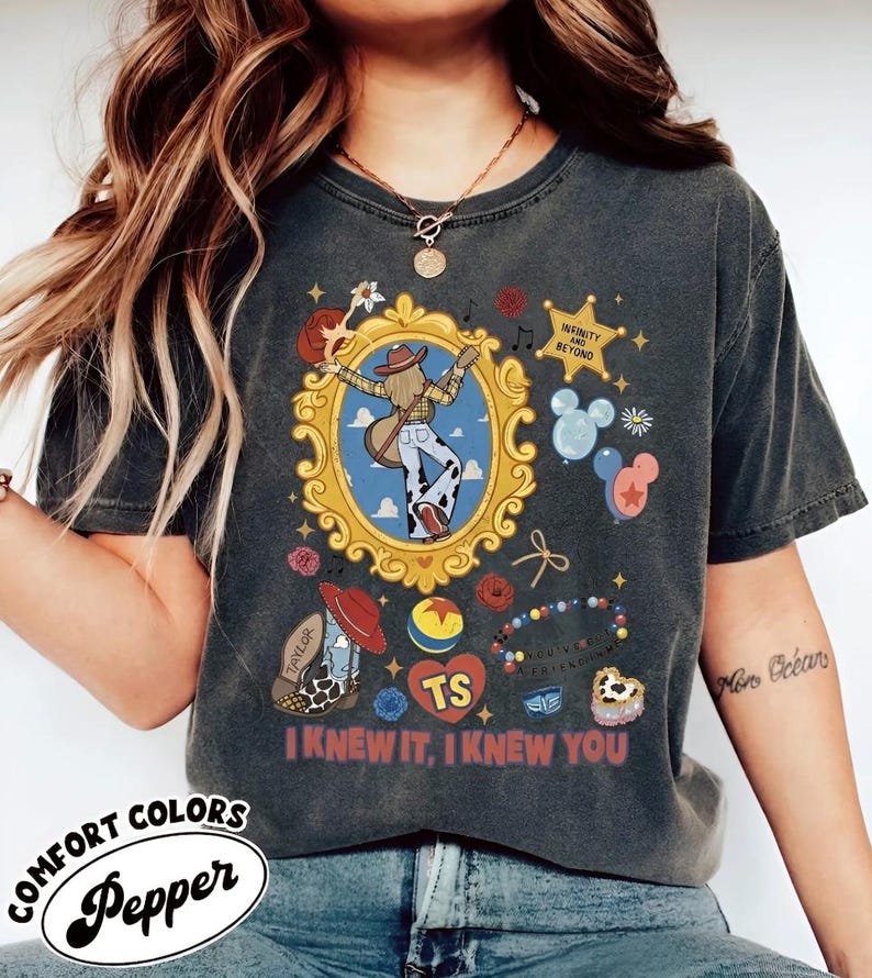 Puede incluir: Una camiseta gris carb&oacute;n con un estampado gr&aacute;fico que presenta un marco dorado ornamentado central con una figura con guitarra, una estrella con el texto INFINITY AND BEYOND, globos, una bota de vaquero, una pelota y el texto I KNEW IT, I KNEW YOU.