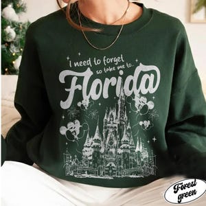 May include: Dark green sweatshirt featuring the phrase "I need to forget so take me to... Florida" in white script. The design includes a castle illustration, fireworks, and Mickey Mouse balloons.