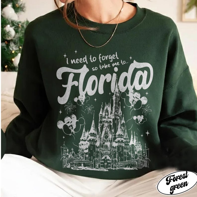 May include: Dark green sweatshirt featuring the phrase "I need to forget so take me to... Florida" in white script. The design includes a castle illustration, fireworks, and Mickey Mouse balloons.