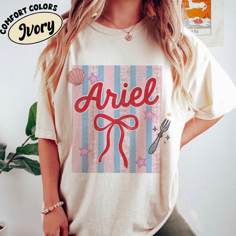 May include: Ivory-colored t-shirt with a graphic featuring the name "Ariel" in red script, a red bow, and a fork illustration. The design includes pink and blue stripes, stars, and a seashell. The shirt has "Comfort Colors" printed on it.