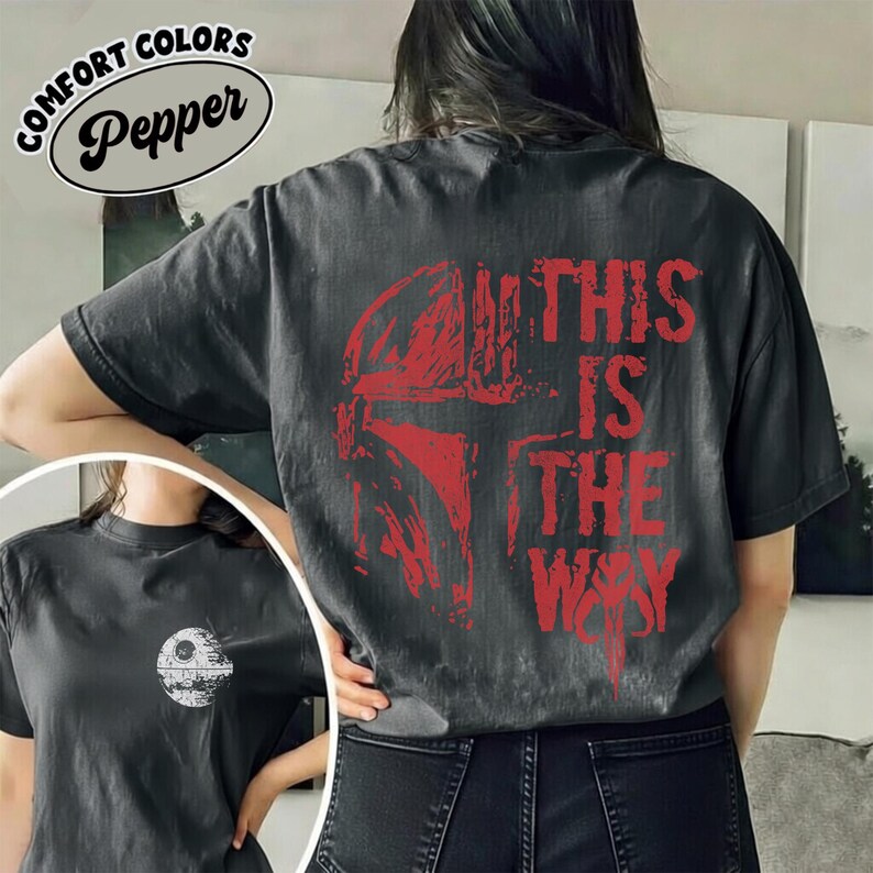 May include: Dark gray t-shirt with a red graphic of a helmet and the text "THIS IS THE WAY." The shirt also features a smaller image of a Death Star. The shirt is part of the "Comfort Colors" collection.