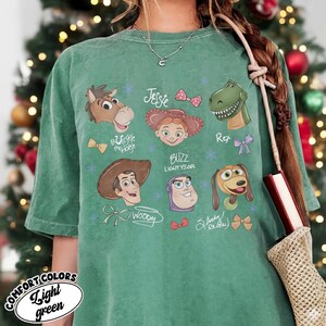 May include: A light green t-shirt featuring cartoon character illustrations from the movie Toy Story. The shirt includes images of Woody, Buzz Lightyear, Jessie, Rex, Slinky Dog, and Bullseye. The text "COMFORT COLORS Light green" is printed on the bottom left.
