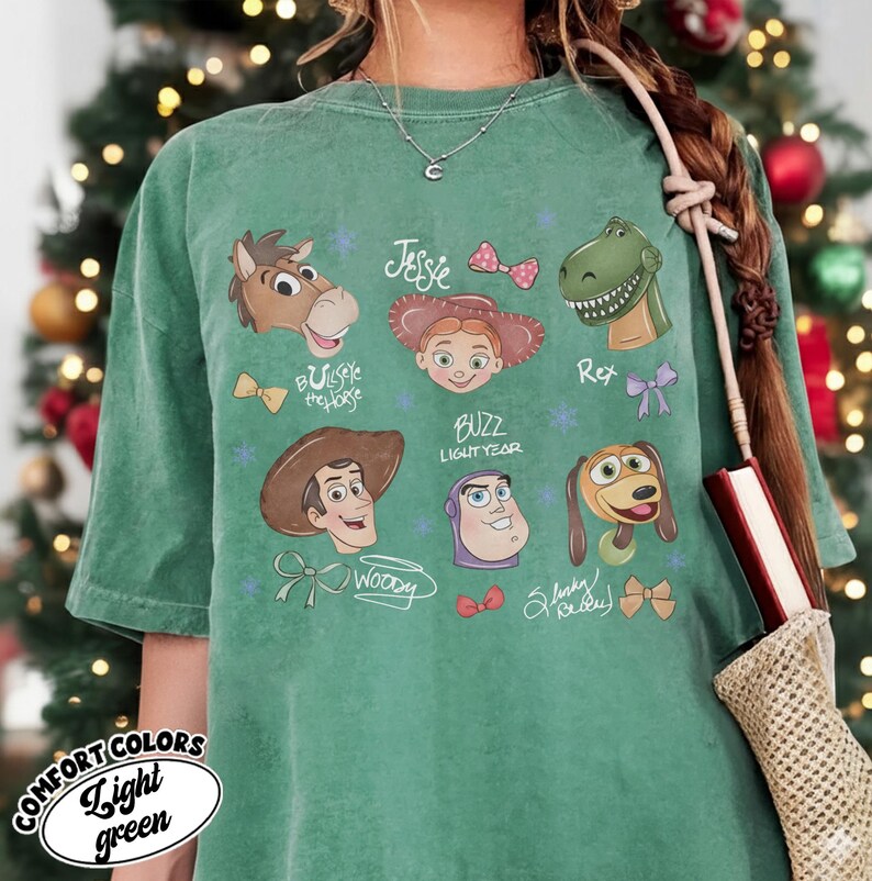 May include: A light green t-shirt featuring cartoon character illustrations from the movie Toy Story. The shirt includes images of Woody, Buzz Lightyear, Jessie, Rex, Slinky Dog, and Bullseye. The text "COMFORT COLORS Light green" is printed on the bottom left.