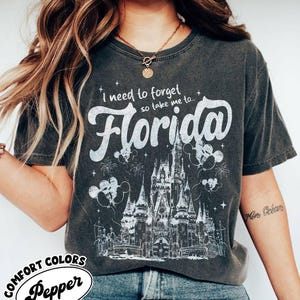 May include: Dark grey Comfort Colors t-shirt featuring the text "I need to forget so take me to... Florida" in white script. The design includes a castle illustration, fireworks, and Mickey Mouse balloons. The shirt is labelled "Pepper".