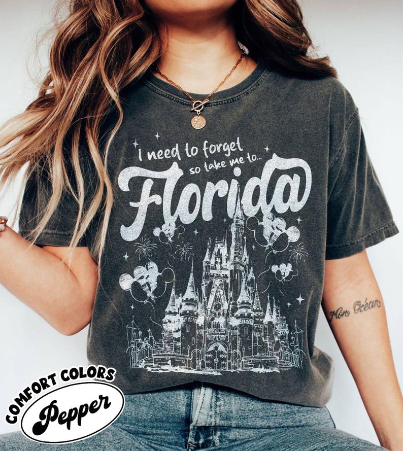 May include: Dark grey Comfort Colors t-shirt featuring the text "I need to forget so take me to... Florida" in white script. The design includes a castle illustration, fireworks, and Mickey Mouse balloons. The shirt is labelled "Pepper".