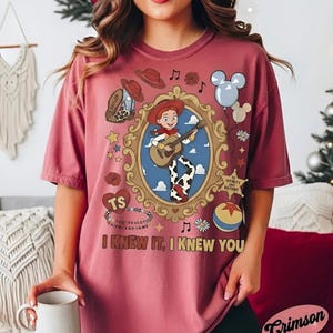 May include: A dusty rose t-shirt featuring a graphic of a cowgirl character playing a guitar inside an ornate gold frame. The design includes a cowboy boot, a sheriff badge with text, a colorful ball, balloons, and the text I KNEW IT, I KNEW YOU.