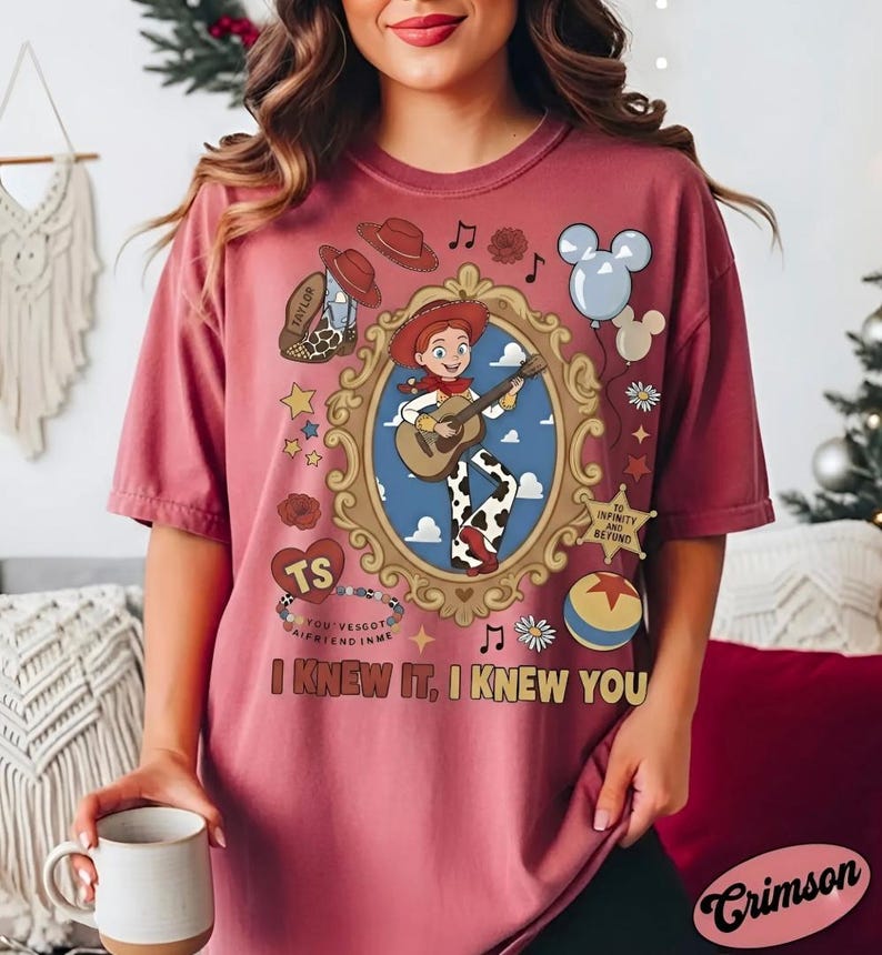 May include: A dusty rose t-shirt featuring a graphic of a cowgirl character playing a guitar inside an ornate gold frame. The design includes a cowboy boot, a sheriff badge with text, a colorful ball, balloons, and the text I KNEW IT, I KNEW YOU.