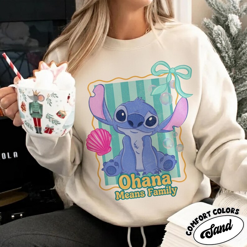 May include: Sand-colored sweatshirt featuring a blue cartoon character with large ears and the text "Ohana Means Family." The graphic includes a pink seashell, a blue bow, and bubbles. The sweatshirt is made of Comfort Colors material.