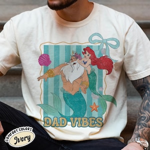 May include: Ivory-colored t-shirt featuring a cartoon illustration of King Triton and Ariel from The Little Mermaid, with the text "DAD VIBES." The design includes a seashell, starfish, and a blue bow. The shirt is labeled "Comfort Colors Ivory."