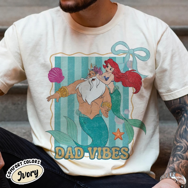 May include: Ivory-colored t-shirt featuring a cartoon illustration of King Triton and Ariel from The Little Mermaid, with the text "DAD VIBES." The design includes a seashell, starfish, and a blue bow. The shirt is labeled "Comfort Colors Ivory."