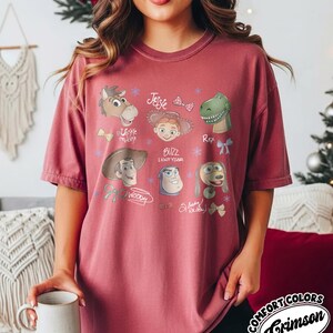 May include: Crimson Comfort Colors t-shirt featuring cartoon illustrations of Toy Story characters. The design includes Woody, Buzz Lightyear, Jessie, Rex, and others. The shirt has a relaxed fit and short sleeves.