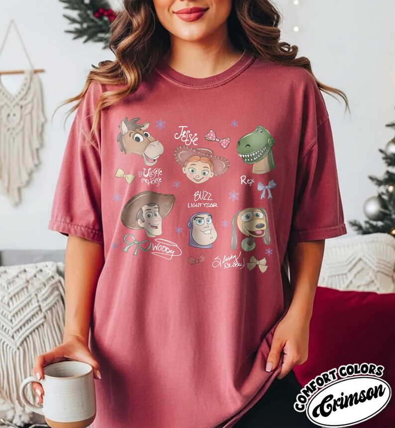 May include: Crimson Comfort Colors t-shirt featuring cartoon illustrations of Toy Story characters. The design includes Woody, Buzz Lightyear, Jessie, Rex, and others. The shirt has a relaxed fit and short sleeves.
