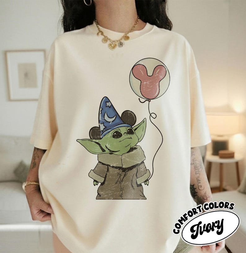 May include: Ivory-colored t-shirt featuring a green alien character wearing a blue wizard hat with a crescent moon and stars. The character is holding a pink Mickey Mouse balloon. The shirt has the words "COMFORT COLORS" and "Ivory" printed on it.