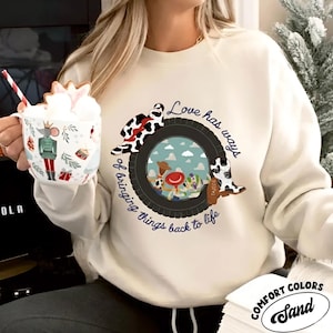 Puede incluir: Una sudadera de cuello redondo color crema con un gr&aacute;fico de un neum&aacute;tico negro que enmarca un fondo azul cielo con personajes de dibujos animados. El texto dice "Love has ways of bringing things back to life" con un sombrero y una bota con estampado de vaca.