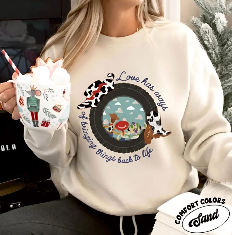 Puede incluir: Una sudadera de cuello redondo color crema con un gr&aacute;fico de un neum&aacute;tico negro que enmarca un fondo azul cielo con personajes de dibujos animados. El texto dice "Love has ways of bringing things back to life" con un sombrero y una bota con estampado de vaca.