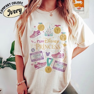 Puede incluir: Camiseta de color marfil con un diseño gráfico con el texto "run Disney Princess". El diseño incluye zapatillas para correr, medallas, un castillo y dorsales de maratón. La camiseta está etiquetada como "Comfort Colors".