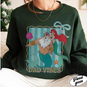 May include: Forest green sweatshirt with a graphic of a merman and mermaid with the text "DAD-VIBES". The graphic is in a rectangular frame with a blue striped background. A seashell, starfish, and bow are also included in the design.