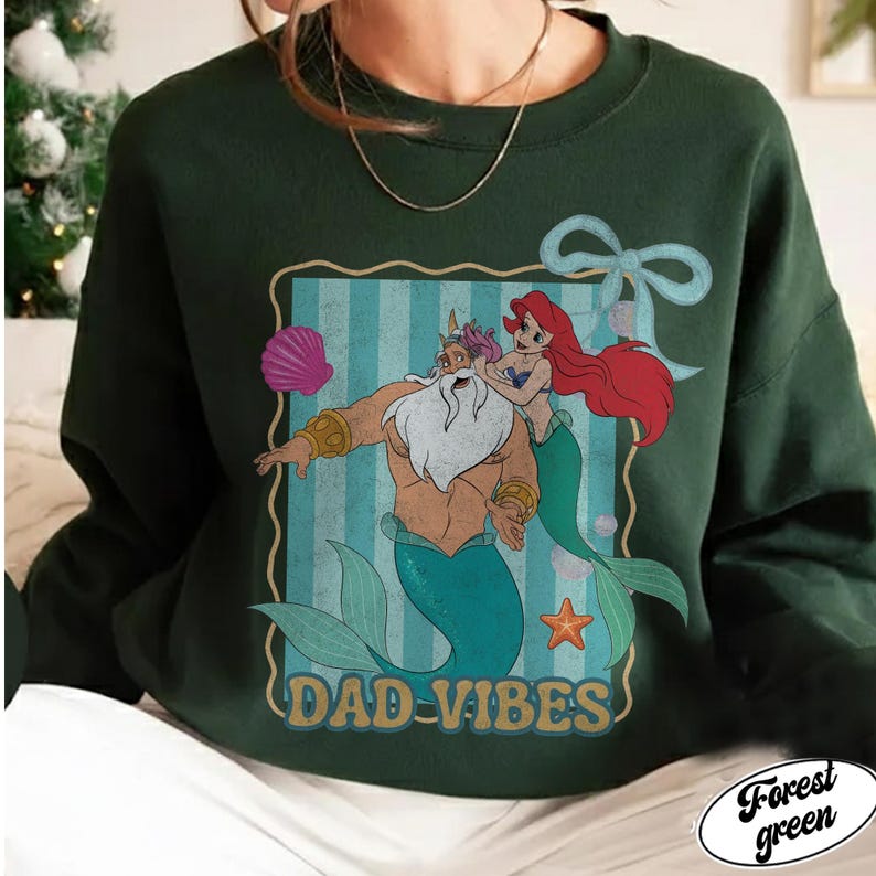 May include: Forest green sweatshirt with a graphic of a merman and mermaid with the text "DAD-VIBES". The graphic is in a rectangular frame with a blue striped background. A seashell, starfish, and bow are also included in the design.