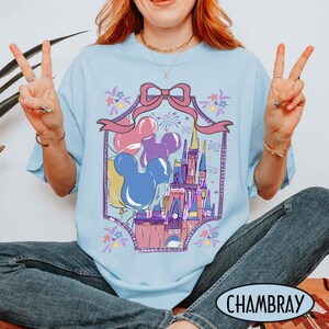 May include: A light blue t-shirt featuring a graphic print of a castle, colorful balloons, and a large pink bow. The design is framed by a decorative border with small floral accents and fireworks, creating a whimsical scene on the front of the shirt.