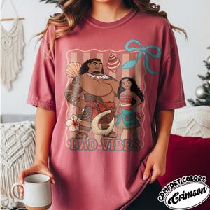May include: Crimson Comfort Colors t-shirt featuring a graphic of two cartoon characters with the text "DAD VIBES". The design includes a large hook, a flower, and a blue bow. The shirt is a solid color.