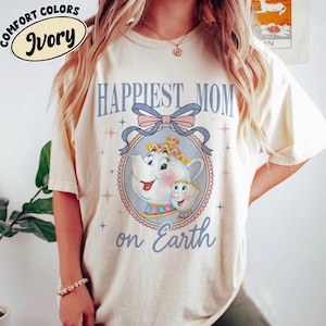 Puede incluir: Camiseta de color marfil con las palabras "Happiest Mom on Earth" en azul claro. La camiseta presenta una ilustración de dibujos animados de una tetera y una taza con un lazo. Las palabras "Comfort Colors" e "Ivory" están en un círculo en la parte superior izquierda.