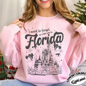 May include: Pale pink sweatshirt featuring the words "I need to forget so take me to... Florida" in black. The design includes a castle, fireworks, and Mickey Mouse balloons. The sweatshirt is labelled "Comfort Colors".